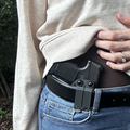 A close-up of a person wearing a black belt holster with a handgun inside, worn over a black belt with blue jeans and a beige sweatshirt.